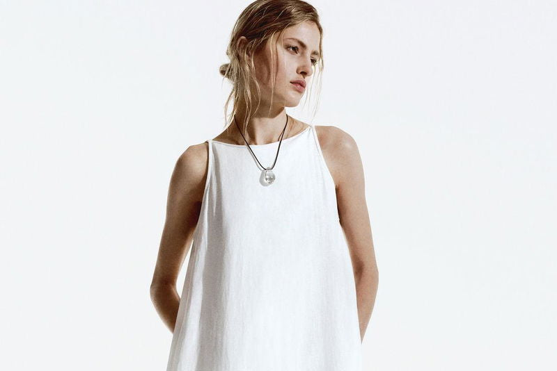 A woman modeling a sleeveless white linen jumpsuit with wide-leg pants and a high neckline, paired with a silver pendant necklace and black sandals, against a plain white studio background.